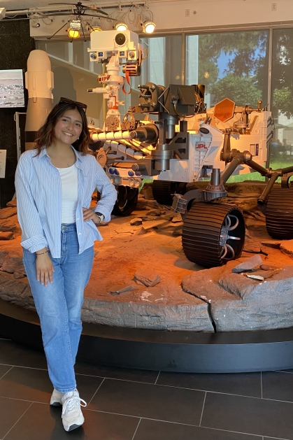 Female young woman poses with her hand on her hip, wearing jeans and a light blue shirt. She has long dark hair and she is standing in front of a lunar rover.