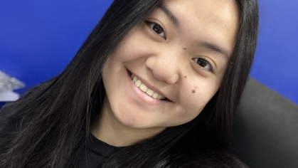 Young woman poses against a blue background. She has black hair and is wearing a black shirt.