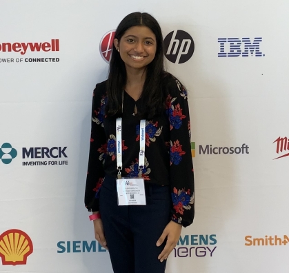 Female engineering student poses in front of a backdrop with corporate logos. She is wearing a black top with red flowers and she has a name badge around her neck.