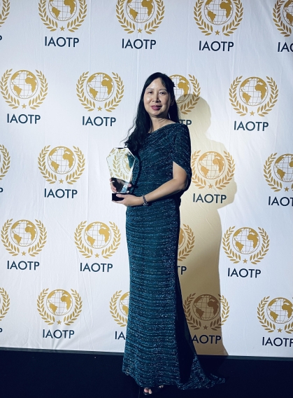 Asian female with long black hair standing in front of an IAOTP step and repeat banner in a black long gown holding an award. made of glass