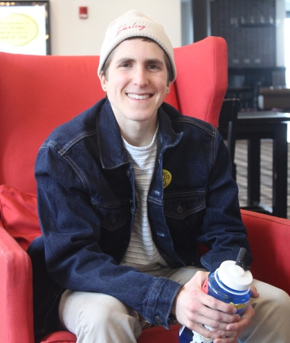 White college student sits in a red wing chair. He is wearing a beanie cap and a dark blue jacket, holding a coffee cup.