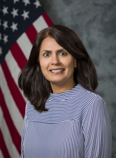 Headshot of woman with shoulder length brown hair in a striped blue blouse with the Aemrican flag behind her.