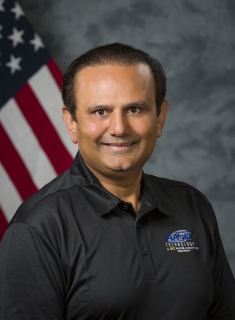 Headshot of s smiling man wearing a navy blue polo shirt with the American flag behind him.