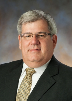 headshot of grey haired man wearing glassed in a dark suit and gold tie