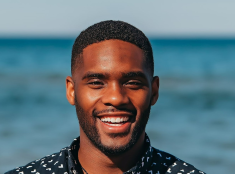 Headshot of Black males student smiling against a blue background. He's wearing a black and white shirt.