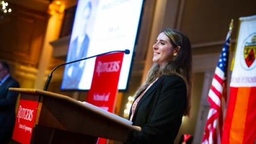 Side view of a young woman delivering remarks from a podium .Behind her is a large projection screen, American flag, and al orange and white banner.