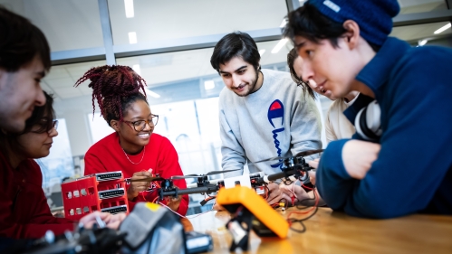 A group of six students, male and female gathered around a wooden table working on a drone.