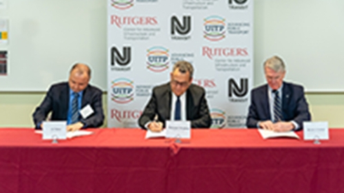 Three men sit at a table draped with a red table cloth each signing a document.