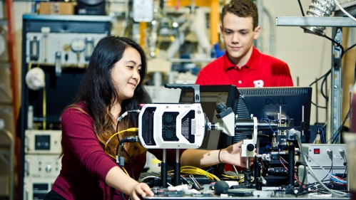 An Asian female student with long black hair, wearing maroon long sleeve shirt and a male white student wearing safety goggles and a red polo shirt conducting research in an engineering lab.