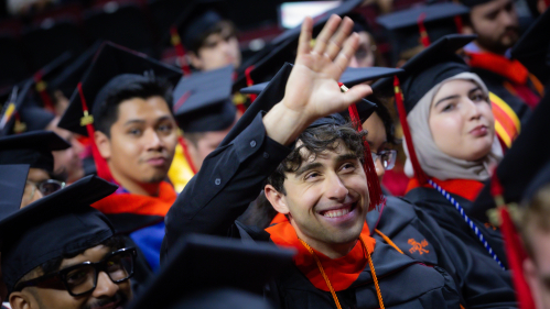 A male college student waves to the crowd at graduation surrounded by his fellow graduates. 