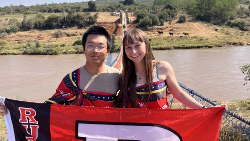 2 students stand on a bridge overlooking river, holding a Rutgers "R" flag together