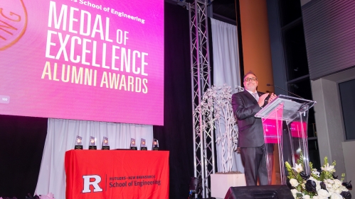 Man standing on a stage behind a podium. The stage includes a table with a red tablecloth with a Rutgers logo.