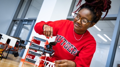 Black female student with braid bun on top of her head, wearing glasses and a red Rutgers sweatshirt is working on a table with drone.