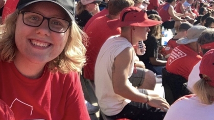 Girl with blond hair and glasses wearing a Rutgers baseball cap and Rutgers T-shirt sitting in the football stadium during a game.