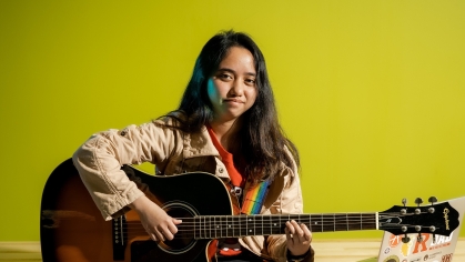 Young woman with long dark hair sits on table holding a guitar.