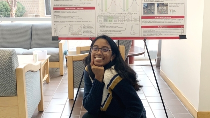 Young woman with black hair and glasses is crouching with hand under chin in front of a research poster.