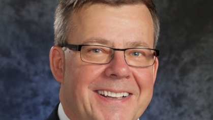 Headshot of white male with grey hair, glasses, wearing a dark suit, white shirt and red tie.