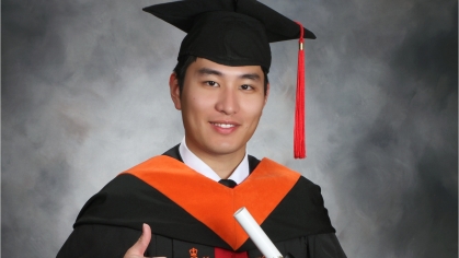 Male student of Chinese dissent wearing a graduation cap and gown holding a diploma and giving a thumbs up sign.