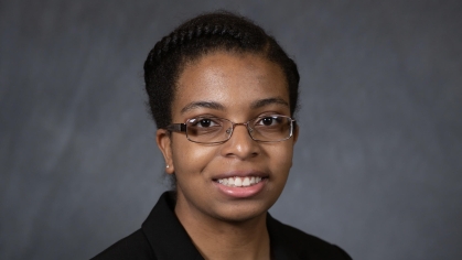 Head shot of young black college student. She is wearing a black suit jacket with a white top underneath. Her hair is short and she has glasses.