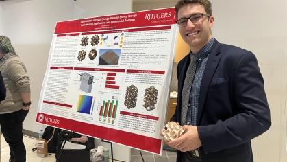 White male university student poses beside a research poster. He has light brown curly hair and is wearing a blue blazer, lighter blue shirt and tie. He is smiling at the camera.