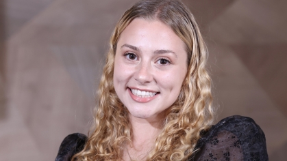 Young white woman with blond curly hair and brown eyes smiles at camera. She is wearing a black top with puffy sheer sleeves.
