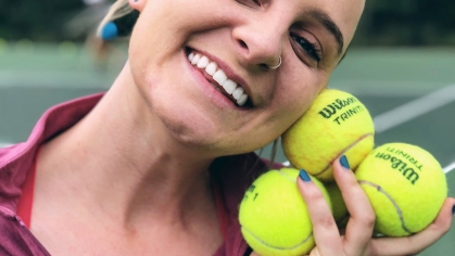 Young white woman poses on a tennis court in a headshot that includes her holding three tennis balls close to her face. She's wearing a pink top.