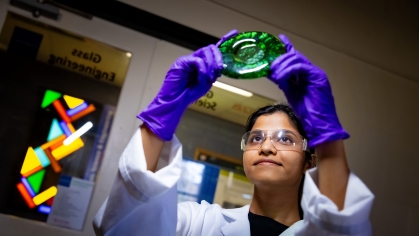 Woman wearing a lab coat and purple safety gloves holds up a round piece of green glass and looks at it.