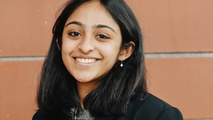 Young woman poses in front of a brick wall. She has black hair and is wearing a black jacket.