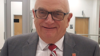 Headshot of bald male with eyeglasses wearing a grey suit, shite button down shirt, and red patterned tie.