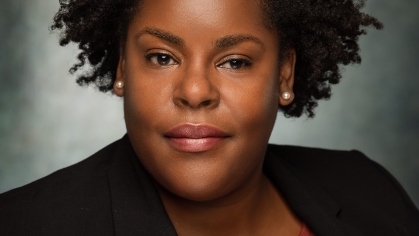 Headshot of Black woman with short dark hair and pearl earrrings, wearing a black suit jacket with a brown sweater.