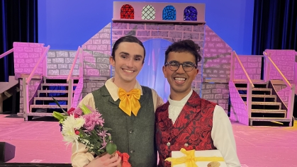 Two young men wearing period costumes pose on a stage holding flowers and a gift. The backdrop is a play setting.