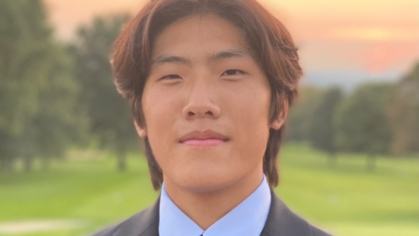 Asian male college student poses outdoors for a headshot. He is wearing a dark suit, light blue shirt and blue tie.