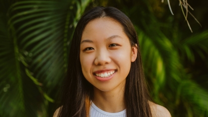 Female college student poses outside in front pine trees. She has long dark hair and is wearing a white sleeveless top.