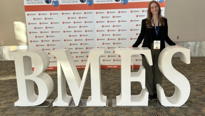 Female college student stands in front of oversized display letters BMES smiling. She is wearing black blazer and dark pants.