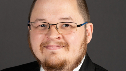 Headshot of male with a buzz haircut, facial hair, wearing a black suit, with a white shirt, and a dark tie.