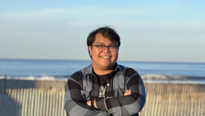 Male student stands wearing a black and grey plaid button down shirt, black horn rimmed eyeglasses with his arms folded with the ocean in the background. 