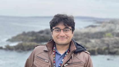 Male engineering student headshot posing outdoors with water and a rocky coastline behind him. He has dark hair, glasses and is wearing a brown jacket over a blue shirt.