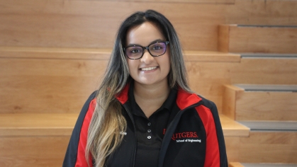 Young female college student with long brown hair parted in the middle with glasses, poses wearing a black and red zip-up jacket.