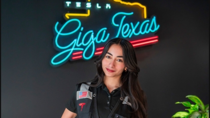 Female engineering student poses wearing a work vest and holding under her arm a hard hat. She is standing in front of the neon sign that says Giga Texas. She has long black hair.