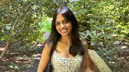 Student poses outdoors with trees in the background. She has long black hair and is wearing a yellow sundress.