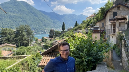 Young male wearing eyeglasses and a blue button down patterned shirt stands with a mountain, a lake, and stone and terra cotta houses in the background. 