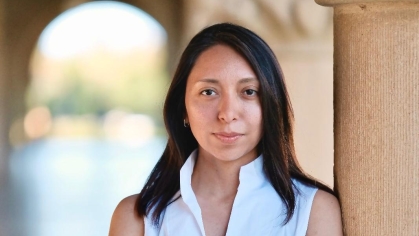 Young woman with long black hair wearing a sleeveless white blouse with her arms crossed leans against a column.