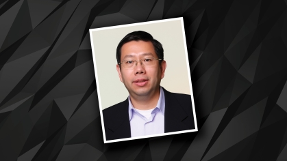 Headshot placed on black and grey patterned background of male with eyeglasses wearing a black suit jacket and white button down shirt.