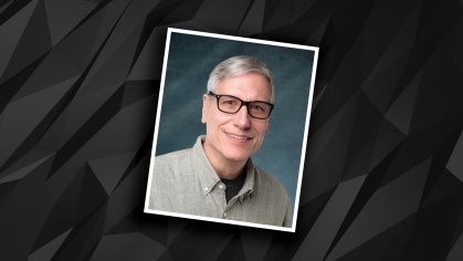 Headshot of male with short grey hair and eyeglasses wearing a grey button down shirt against a grey and black patterned background.