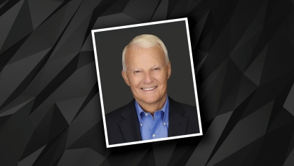 Headshot of male with short white hair wearing a blue button down shirt and a black suit jacket against a black and grey patterned background