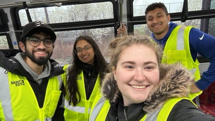 Four students pose on a bus wearing yellow safety vests.
