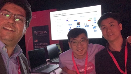 Male professor poses for a closeup with two male students with a monitor display behind them.