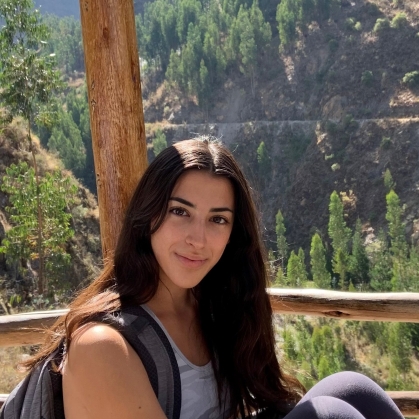 Young woman with long dark hair sitting with a back pack with a view of trees and mountains behind her.