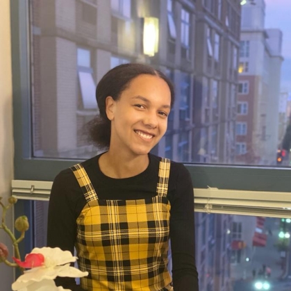 Young Black woman sits on a window sill with a city view behind her and white and red flowers in the foreground. She is wearing a yellow and black plaid jumper with a black shirt.