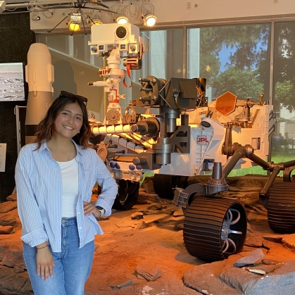 Female young woman poses with her hand on her hip, wearing jeans and a light blue shirt. She has long dark hair and she is standing in front of a lunar rover.